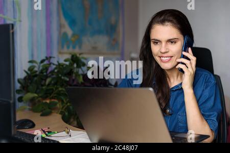 Überglücklich junge Frau im Denim-Shirt arbeitet zu Hause mit einem Laptop, spricht per Telefon und schaut auf den Laptop. Weltkarte im Hintergrund Stockfoto