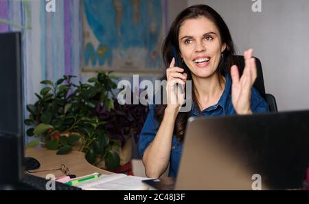 Überglücklich junge Frau im Denim-Shirt arbeitet zu Hause mit einem Laptop, spricht per Telefon und schaut nach oben. Weltkarte im Hintergrund Stockfoto