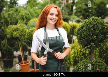 Schöne Dame mit rothaarigen lockigen Haaren in Schürze stehen und halten große Gartenschere, während glücklich in der Kamera mit Pflanzen auf dem Hintergrund suchen Stockfoto