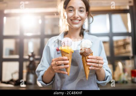 Weibliche fröhliche Verkäuferin hält zwei leckere Eis in Waffelkegel drinnen, Verkauf von Süßigkeiten im Shop Stockfoto