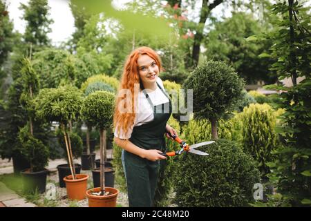 Junge Gärtnerin mit rothaarigen Lockenhaaren, die in der Schürze steht und große Gartenscheren hält, während sie freudig in der Kamera im Freien schaut Stockfoto