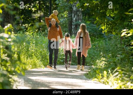 Junge Familie von Eltern und zwei kleinen Kindern, die im Wald spazieren - Mama und Papa halten ihre Tochter auf Skate-Rollen an den Händen. Mittlerer Schuss Stockfoto