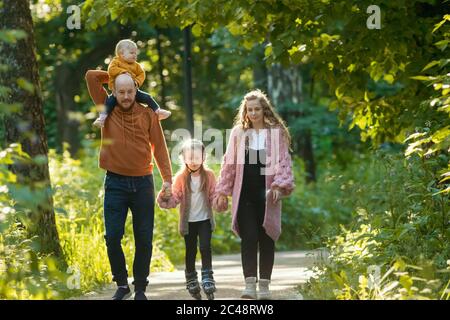 Junge Familie von Eltern und zwei Kindern, die im Park spazieren - Mama und Papa halten ihre Tochter auf Skate-Rollen an den Händen. Mittlerer Schuss Stockfoto