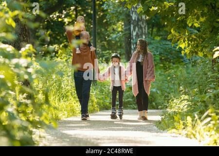 Junge Familie von Eltern und zwei kleinen Kindern, die im Park spazieren - Mama und Papa halten ihre Tochter auf Skate-Rollen an den Händen. Mittlerer Schuss Stockfoto