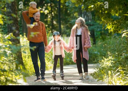 Junge glückliche Familie von Eltern und zwei Kindern, die im Park spazieren - Mama und Papa halten ihre Tochter auf Skate-Rollen an den Händen. Mittlerer Schuss Stockfoto