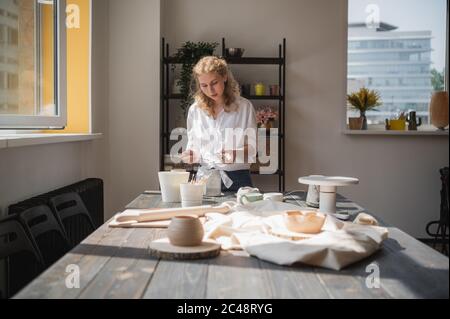 Eine junge europäische schöne Töpferin in einem kreativen Prozess der Herstellung von Keramik am Tisch in der Werkstatt. Haare flattern im Wind an sonnigen Tag Stockfoto
