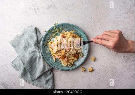 Die Hand der Frau hält eine Gabel mit Pasta Fettuccine mit Pilzen, Speck, Parmesan und Bechamelsauce in Teller auf hellem Holzhintergrund, Draufsicht Stockfoto