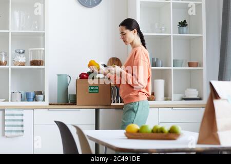 Junge Frau, die auf ihr Handy schaut und die Lebensmittel in der Box prüft, während sie in der Küche steht Stockfoto