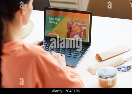 Rückansicht einer Frau, die vor dem Laptop am Tisch sitzt und online Essen bestellt Stockfoto