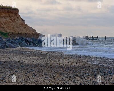 Nordseewellen brechen gegen Felsen und schützen weiche Sandklippen vor Erosion. Reste eines Anlegesteges führen ins Meer und ein Schiff ist nahe an Land. Stockfoto