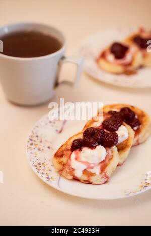 Hausgemachte Käsekuchen aus Hüttenkäse mit saurer Sahne und Himbeermarmelade. Eine Tasse Tee im Hintergrund Stockfoto