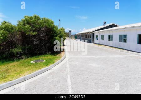 Robben Island, Südafrika - 24. November 2019: Blick auf die Gefängnisgebäude rechts und einen Wachturm am Ende einer gepflasterten Straße auf der Robben Isl Stockfoto