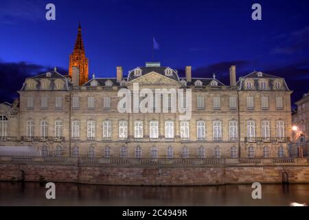 Die Fassade des Rohan-Palastes in Straßburg, Frankreich bei Nacht. Der Turm der Kathedrale Notre-Dame blickt auf das Gebäude. Stockfoto