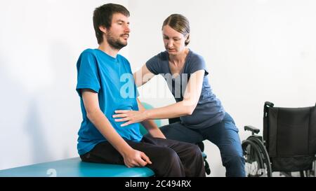 Physiotherapeut, der mit behinderten Menschen im Rollstuhl auf einem Therapietisch trainiert. Stockfoto