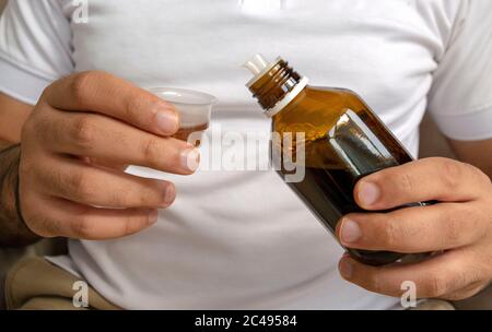 Junger Mann in weißem T-Shirt gießt medizinischen Sirup in Messbecher. Junger Mann hält Flasche Sirup mit Tasse. Stockfoto