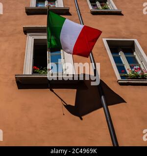 Nahaufnahme einer italienischen Flagge auf einer Fensterbank mit roten und weißen Blumen.die Fassade des alten Gebäudes ist ockerfarben gestrichen. Stockfoto