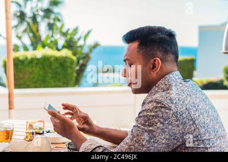Junger, hübscher Inder, der im Resort mit dem Smartphone im Café sitzt Stockfoto