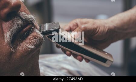 Nahaufnahme des elektrischen Rasierers in den Händen eines Barbiers.Serious Master Schneiden Bart des gealterten Kunden im Salon. Stockfoto