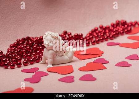 Kleine weiße Gipsstatuette von Amor unter Valentinstag Dekorationen. Herzen und rote Perlen auf pastellrosa Hintergrund verstreut. Liebe und Urlaubskonzept. Stockfoto