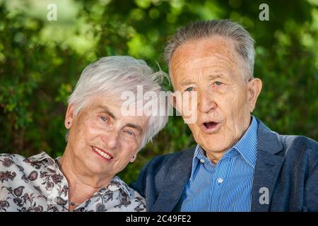 Kopf-Schulter-Porträt eines älteren Rentnerpaares vor grünen Blättern im Sommer, das glücklich auf die Kamera schaut. Mann mit offenem Mund. Stockfoto