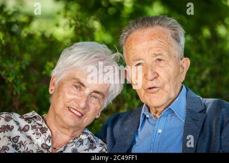 Kopf-Schulter-Porträt eines älteren Rentnerpaares vor grünen Blättern im Sommer, das glücklich auf die Kamera schaut. Stockfoto