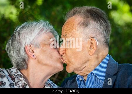 Kopf-Schultern-Porträt eines älteren älteren Ehepaares, das sich im Sommer vor grünen Blättern küsst. Stockfoto