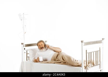 Älterer alter Mann erholt sich in einem bequemen Krankenhausbett isoliert auf weißem Hintergrund. Behandlung erhalten. Konzept der Gesundheitsversorgung und Medizin. Liegen, Telefonieren, Tee trinken. Copyspace. Stockfoto