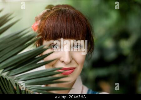 Nahaufnahme Beauty Portrait von Gesicht ziemlich charmante rothaarige Frau, posiert vor der Kamera mit Palmenblatt in der Nähe ihres Gesichts, während im Gewächshaus stehen Stockfoto