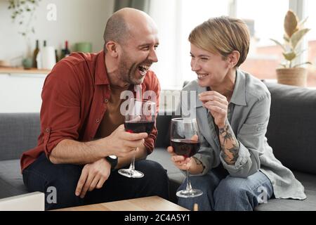Glückliches Paar, das auf dem Sofa mit Weinläsern sitzt und lachend, verbringen sie lustige Zeit zusammen Stockfoto