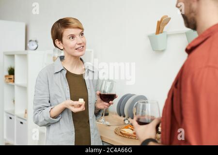 Junge Frau mit kurzen Haaren, die Rotwein trinkt und Käse isst und mit dem Mann spricht, während sie in der Küche stehen Stockfoto