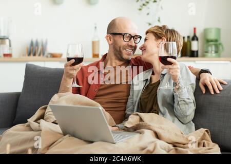 Glückliches reifes Paar, das auf dem Sofa mit Gläsern Rotwein am Laptop sitzt und lacht Stockfoto