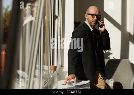 Porträt eines reifen Geschäftsmannes in Brillen, die neben dem Bürofenster stehen und beim Telefonieren auf dem Handy auf die Kamera schauen Stockfoto