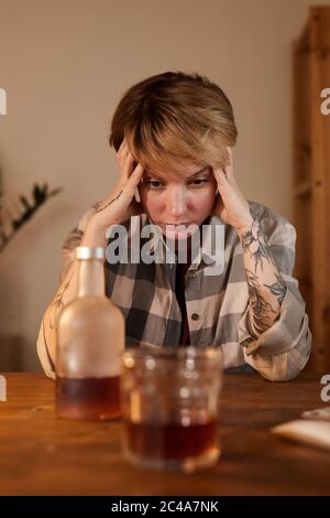 Junge Frau, die am Tisch sitzt und eine Flasche Whisky anschaut, ist betrunken und hat Alkoholsucht Stockfoto