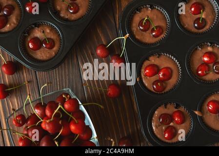 Backen von Schokoladenmuffins. Rohe vegane BananenkirschMuffins mit frischen Beeren auf braunem Holzhintergrund. Draufsicht. Sommeressen Stockfoto