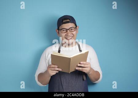 Portrait des asiatischen männlichen Chefkochs, der Buch der Rezepte liest, glücklicher lächelnder Ausdruck, vor blauem Hintergrund Stockfoto