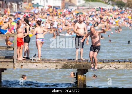 Southend on Sea, Essex, Großbritannien. Juni 2020. Mit dem prognostizierten Rekordtemperaturen für 2020 Menschen sind auf dem Weg an die Küste, um sich abzukühlen, trotz der COVID-19 Coronavirus Beratung. In Southend on Sea ist das Ufer der Themse-Mündung voll, mit Menschen, die schwimmen und ins Meer springen. Weiße kaukasische Männchen springen ins Meer von einem hölzernen Pier mit vollgepackten Strand dahinter Stockfoto
