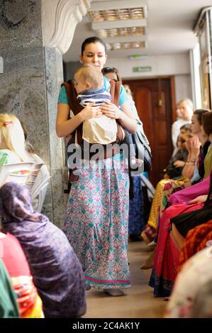 Junge Frau mit Baby auf Händen, die den Korridor des Krishna tempe entlang gehen, Menschen beten Stockfoto