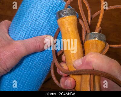 Nahaufnahme der Hände eines Mannes, der eine blau aufgerollte Übungsmatte und ein Holz- und Lederspringseil hält. Stockfoto