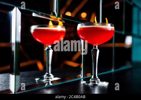 Ein rot-saurer Cocktail in einem Coupé-Glas, garniert mit einer Orangenschale. Spiegelung in einer Glaswand. Stockfoto