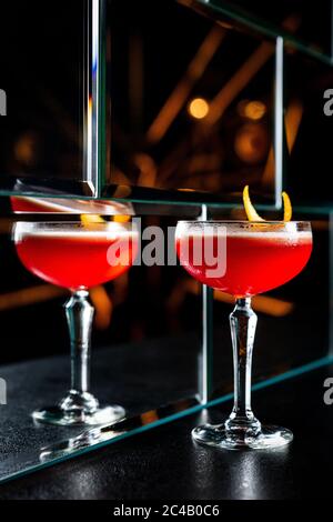 Ein rot-saurer Cocktail in einem Coupé-Glas, garniert mit einer Orangenschale. Spiegelung in einer Glaswand. Stockfoto