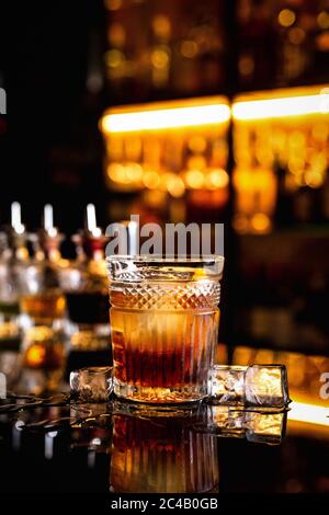 Ein Cocktail in einem altmodischen Glas auf einer Theke mit Spiegelung, Eiswürfel um, Flaschen mit Bittern, Bokeh-Lichter, vertikales Foto Stockfoto