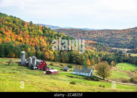 Hügelige ländliche Landschaft mit einem Bauernhof am Fuße eines Gebirges, bedeckt von Laubbäumen auf dem Gipfel des Herbstlaubes an einem bewölkten Herbsttag Stockfoto