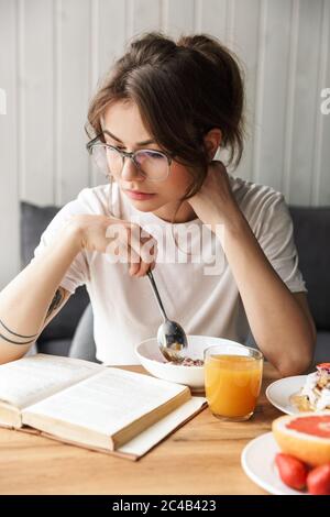 Foto einer jungen denkenden Frau, die Buch liest, während sie zu Hause im gemütlichen Zimmer frühstückt Stockfoto