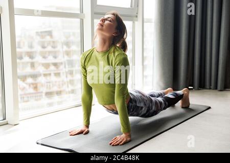 Foto von fokussierten Brünette Frau tun Yoga-Übungen auf Matte beim Training in gemütlichen Raum Stockfoto