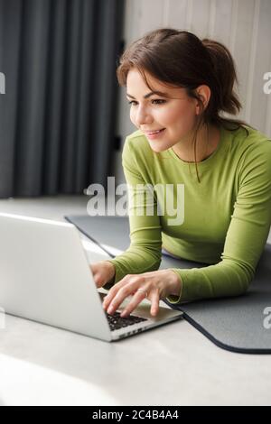 Foto von lächelnden Brünette Sportlerin mit Laptop beim Arbeiten auf Matte in gemütlichen Raum Stockfoto