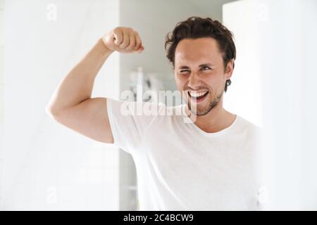 Attraktiver, glücklicher, selbstbewusster junger Mann, der vor dem Badezimmerspiegel steht und Bizeps beugt Stockfoto