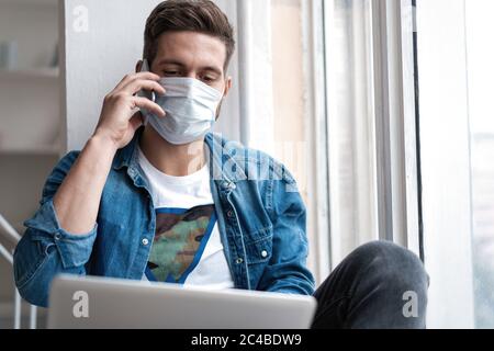 Junger glücklicher Mann sitzt in Schutzmaske am Telefon reden. Stockfoto