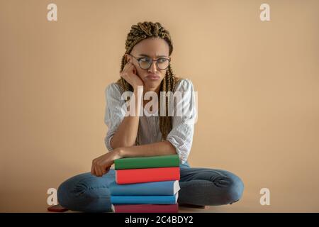 Junge afroamerikanische Frau in Brillen, die auf dem Boden in Lotuspose vor Büchern sitzt Stockfoto