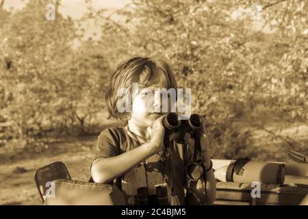 Ein fünfjähriger Junge mit Ferngläsern, der in einem offenen Jeep im Wald steht. Stockfoto