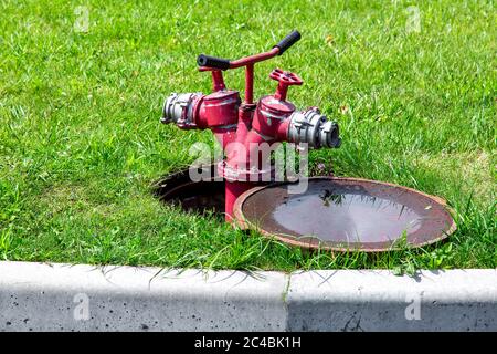 Hydrant, der an ein offenes Mannloch auf einem grünen Rasen mit Gras angeschlossen ist, in der Nähe eines offenen Mannlochs mit einem Schieber. Stockfoto